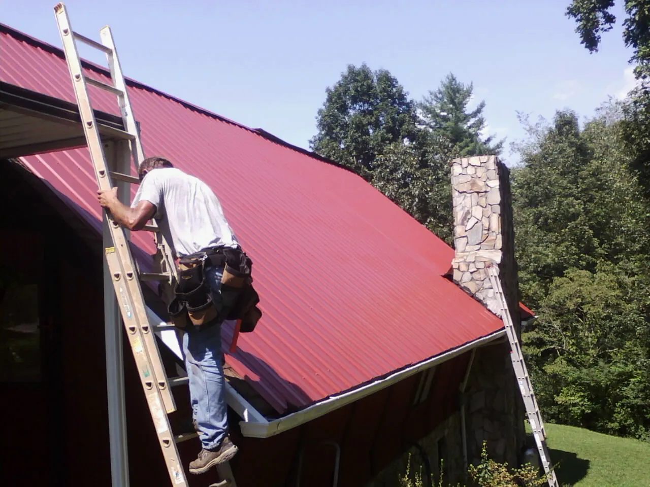 Roofer on a ladder beside a red metal roof and stone chimney — Soffit & Fascia Repair by Roofing Brothers in Perryville